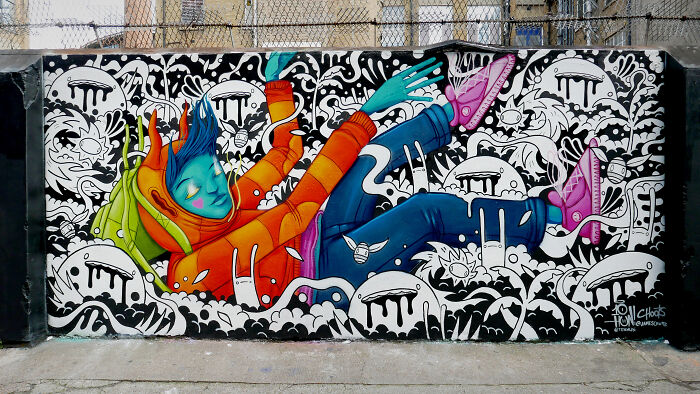 Colorful street art mural of a blue-skinned figure floating among abstract black and white patterns on an urban wall.