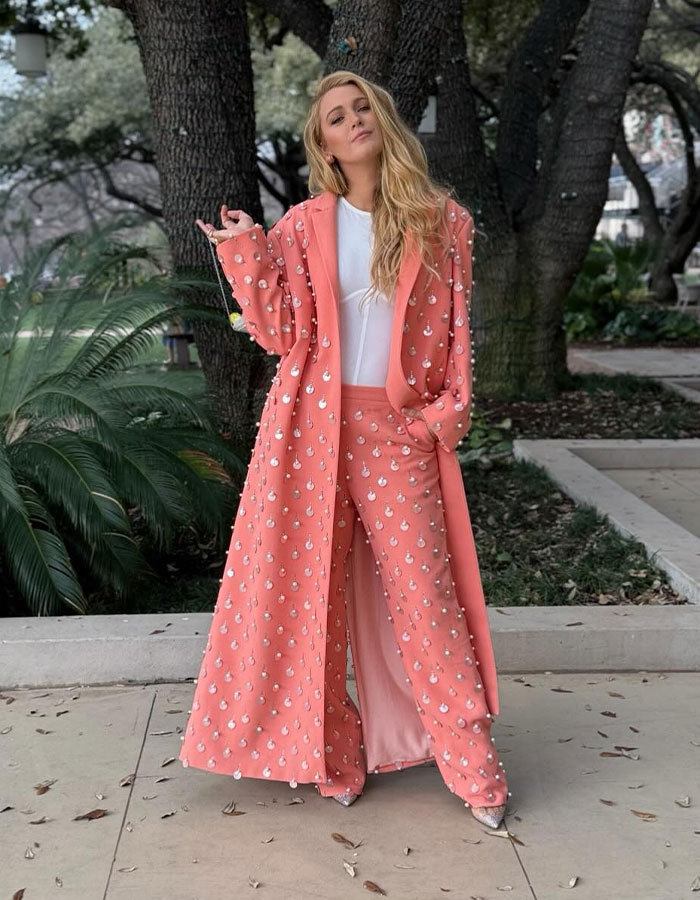 Blonde woman in a coral embellished coat and pants posing outdoors amid trees and greenery in a casual stance.