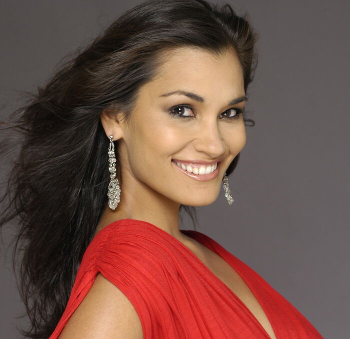 Miss Universe winner smiling confidently in a red dress with flowing hair and sparkling earrings in a studio photo.