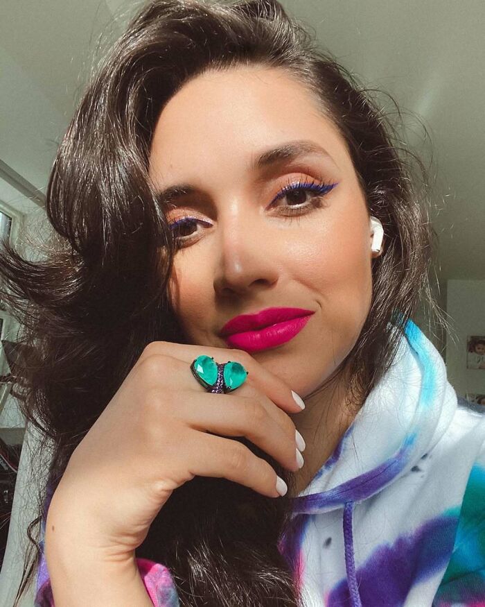Miss Universe winner with bright pink lipstick, blue eyeliner, and a green gemstone ring posing indoors in natural light.