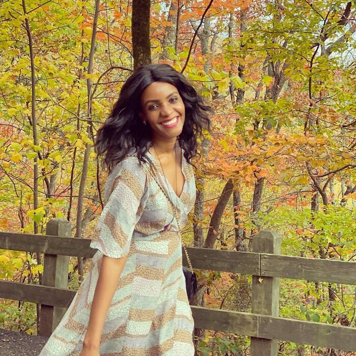 Miss Universe winner posing outdoors in autumn with colorful fall foliage and wooden fence in the background.