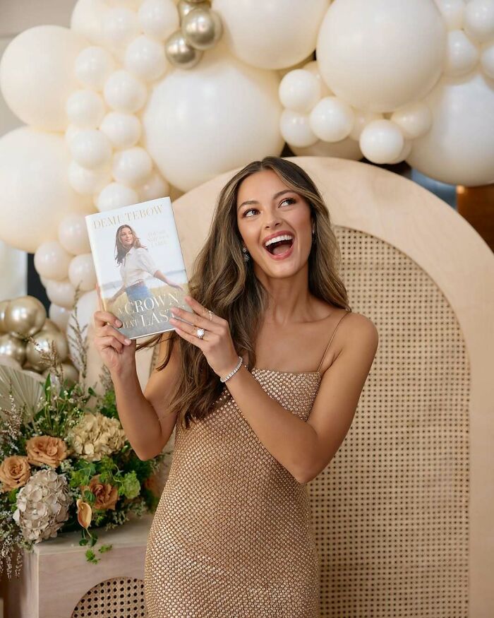 Miss Universe winner smiling and holding a book, standing against a backdrop of cream balloons and floral arrangements.