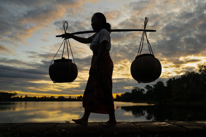 Silhouette of a woman carrying traditional pots by a lakeside at sunset, showcasing the beauty of cultures.