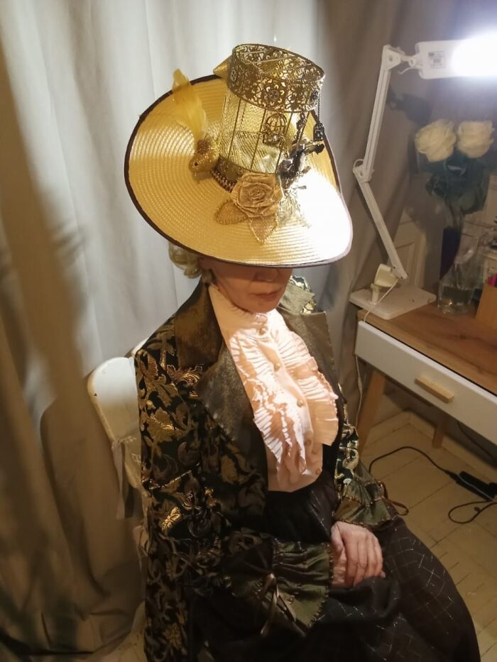 Woman in an incredible cosplay wearing a decorative hat and vintage-style clothing, sitting indoors by a desk with flowers.