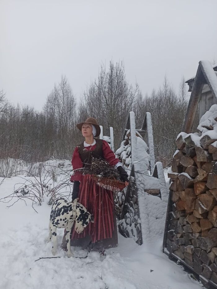 Woman in detailed cosplay outfit standing in snowy forest beside stacked firewood holding a basket and a spotted dog.