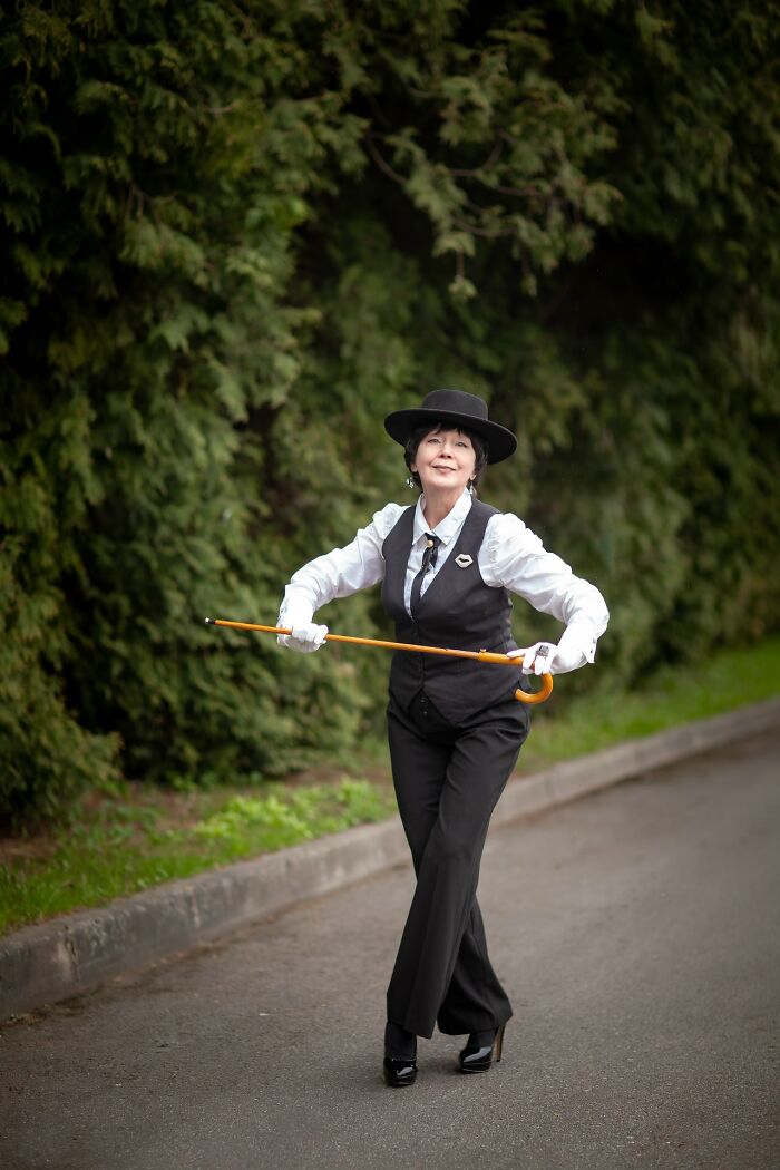 Woman in incredible cosplay outfit with hat and cane posing outdoors on a paved path near greenery.