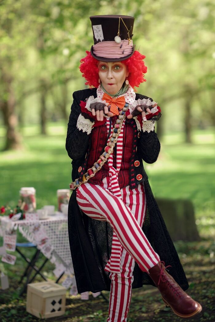 Woman in incredible cosplay dressed as Mad Hatter with red curly hair, striped pants, and detailed costume outdoors in a park.