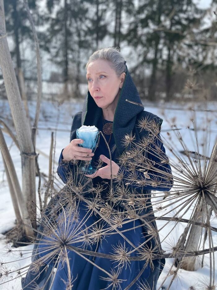 Woman in an intricate cosplay outfit holding a blue cup with foam, posing outdoors in a snowy natural setting.