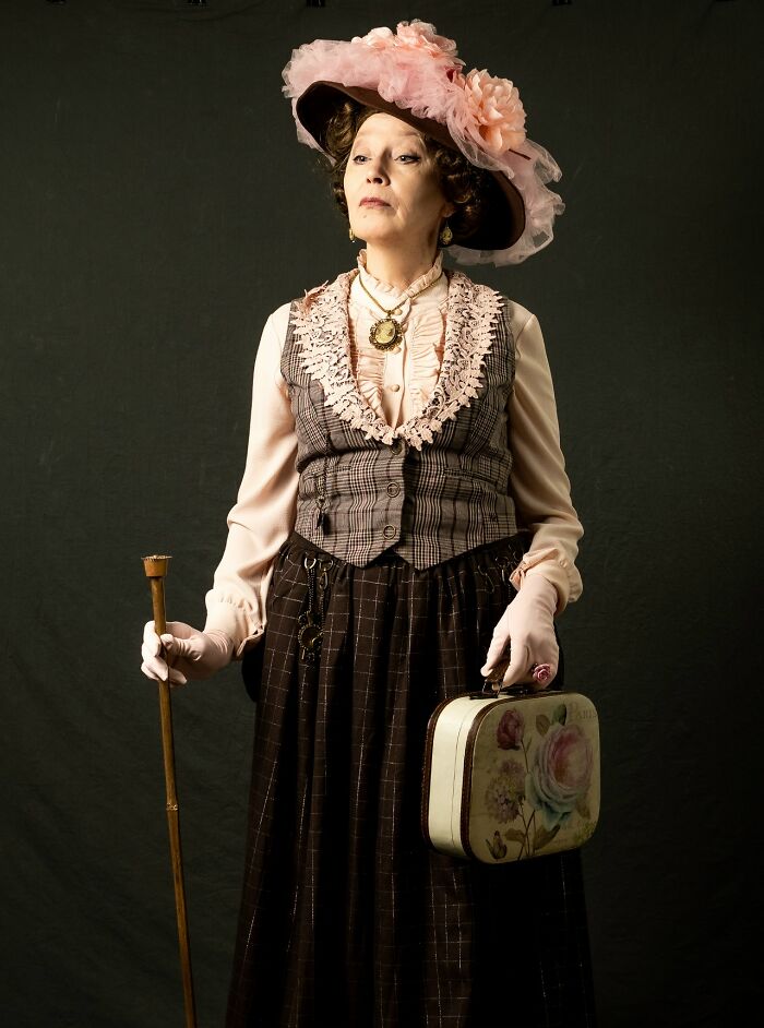 Woman in incredible cosplay wearing vintage outfit with a large pink hat, holding a cane and floral handbag against dark background