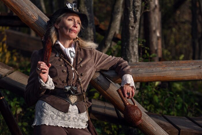 Woman in an incredible cosplay outfit holding a wooden jug, posing outdoors near a rustic wooden fence in a forest setting.
