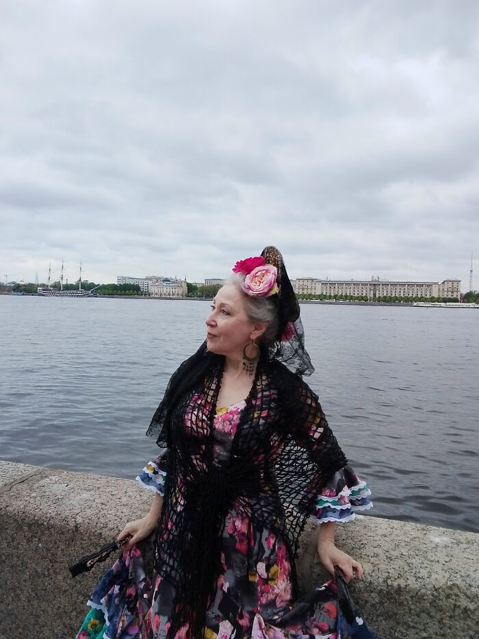 Woman by the water wearing an incredible floral cosplay dress with black lace shawl and flower headpiece outdoors.