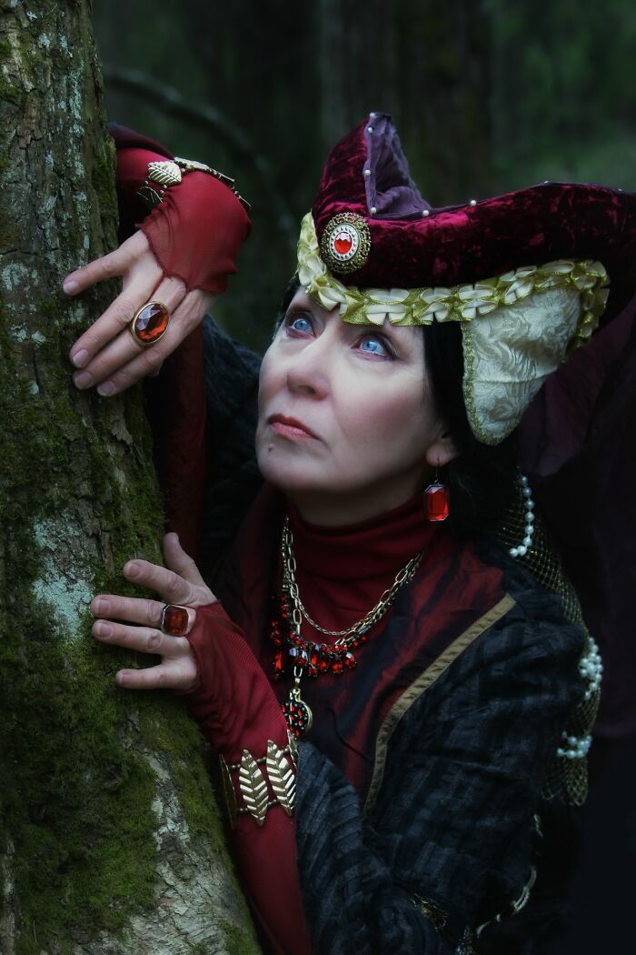 Woman in detailed medieval cosplay costume with jeweled accessories, posing thoughtfully against a tree in a forest setting