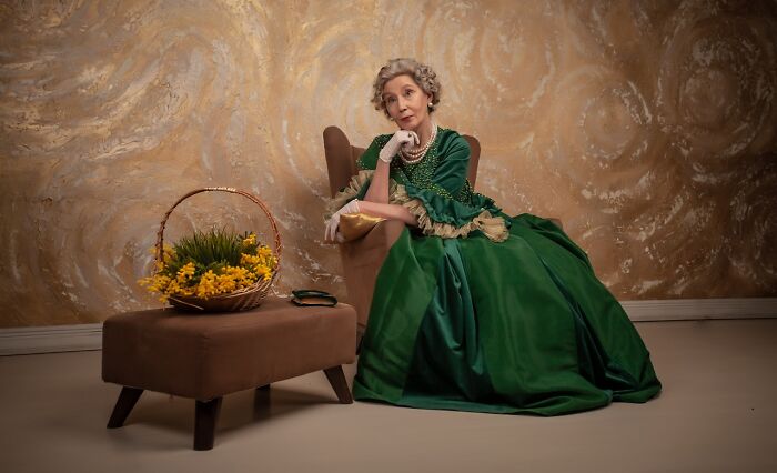 Woman in an elaborate green cosplay dress sitting on the floor beside a brown stool with a basket of yellow flowers