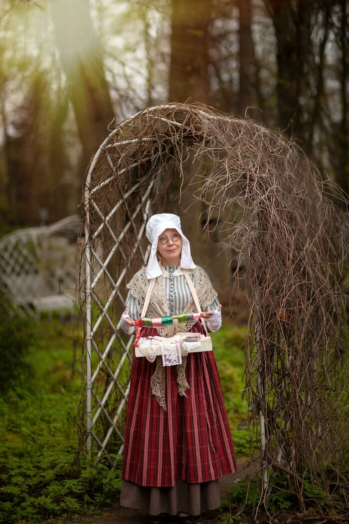 Woman in an incredible cosplay dressed in historical costume, standing under a rustic arch in a forest setting.