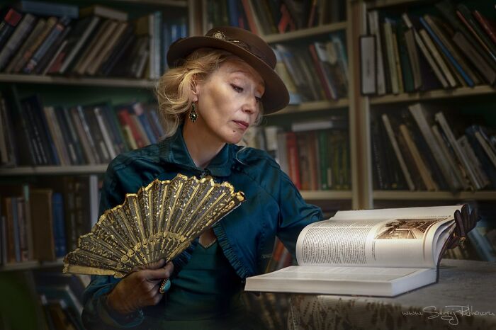 Woman in vintage cosplay outfit with a hat and fan, reading a large book in a library setting, showcasing incredible cosplays.