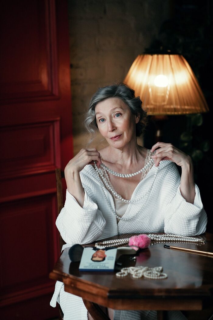 Woman wearing a white robe holding pearl necklaces, sitting at a table with cosplay accessories and makeup tools.