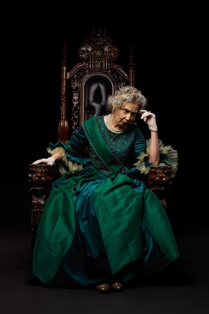 Woman in an incredible cosplay wearing a green royal gown and crown sitting thoughtfully on an ornate throne against a black background