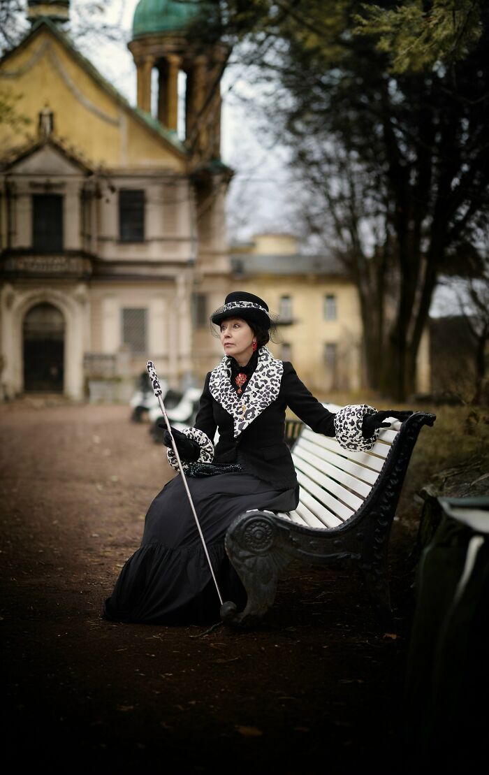 Woman in incredible cosplay with leopard print accents sitting on a park bench holding a cane in front of old building.
