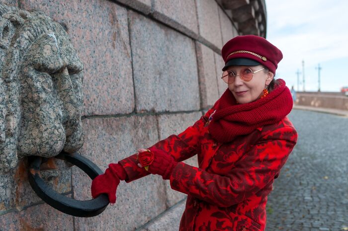 Woman in a red cosplay outfit with scarf and hat posing by a stone wall with a lion head ring sculpture outdoors