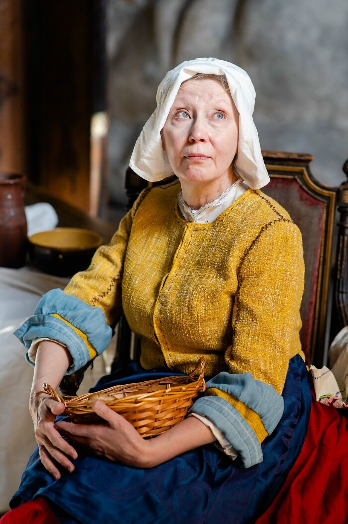 Woman in an incredible cosplay wearing a historical outfit with a white bonnet and holding a wicker basket indoors.