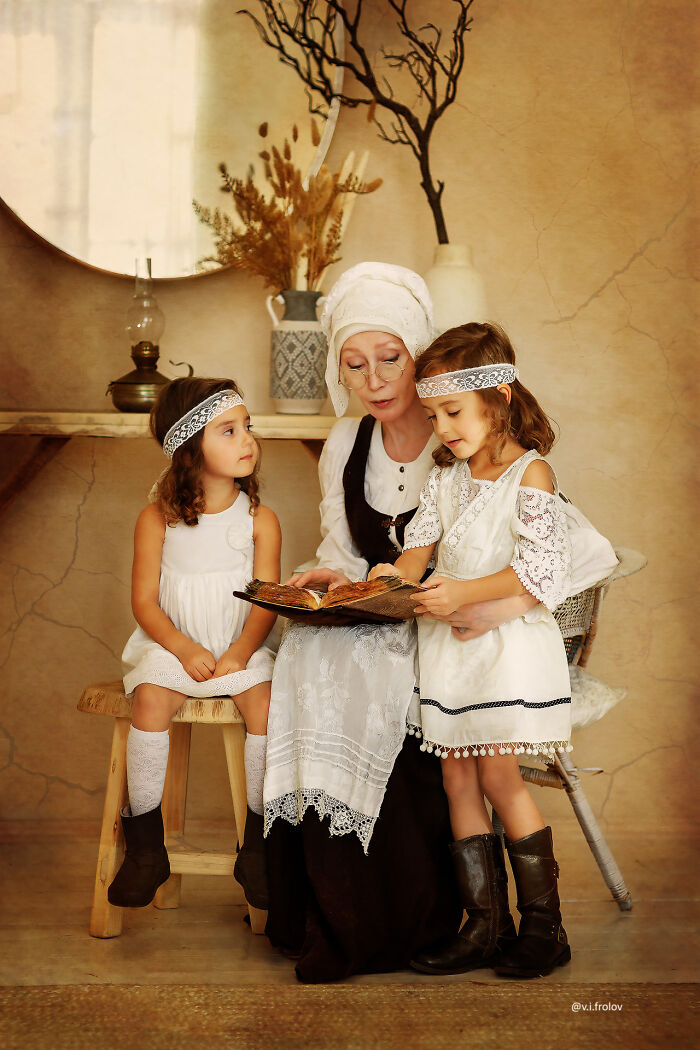 Woman in vintage cosplay reading a book to two girls in white dresses and headbands in a warm rustic setting