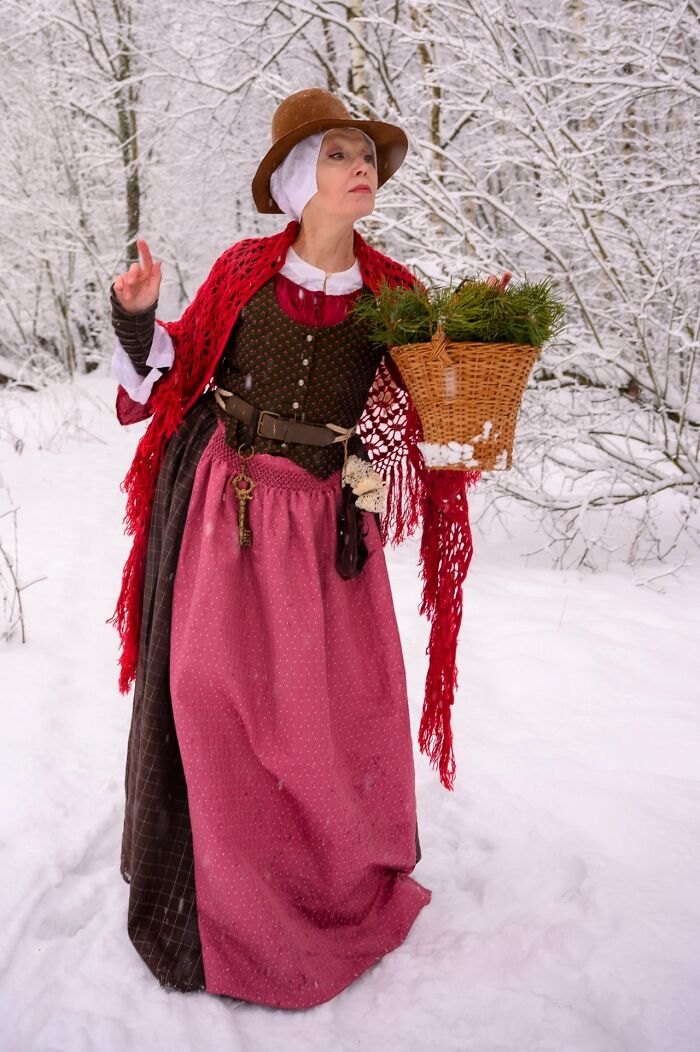 Woman in incredible cosplay wearing vintage dress and red shawl, carrying basket in snowy forest setting.