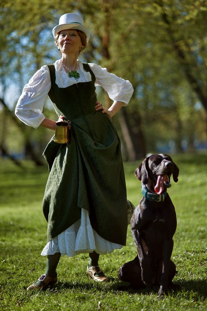 Woman in an incredible cosplay outfit with a green dress and white hat standing outside with a large black dog.