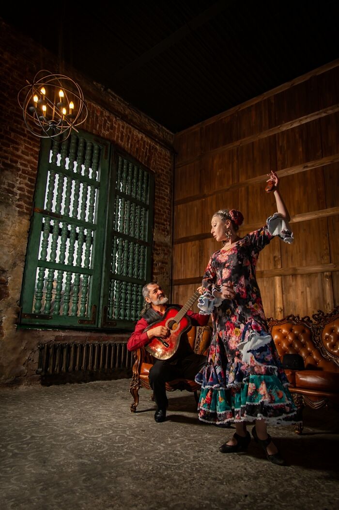 Woman in colorful cosplay dress dancing flamenco while man plays guitar in rustic room setting.