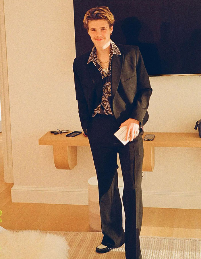 Cruz Beckham dressed in a patterned shirt and black suit, smiling casually indoors, fashion inspired by viral speedo shot. Cruz Beckham dressed in a patterned shirt and black suit, smiling casually indoors, fashion inspired by viral speedo shot.
