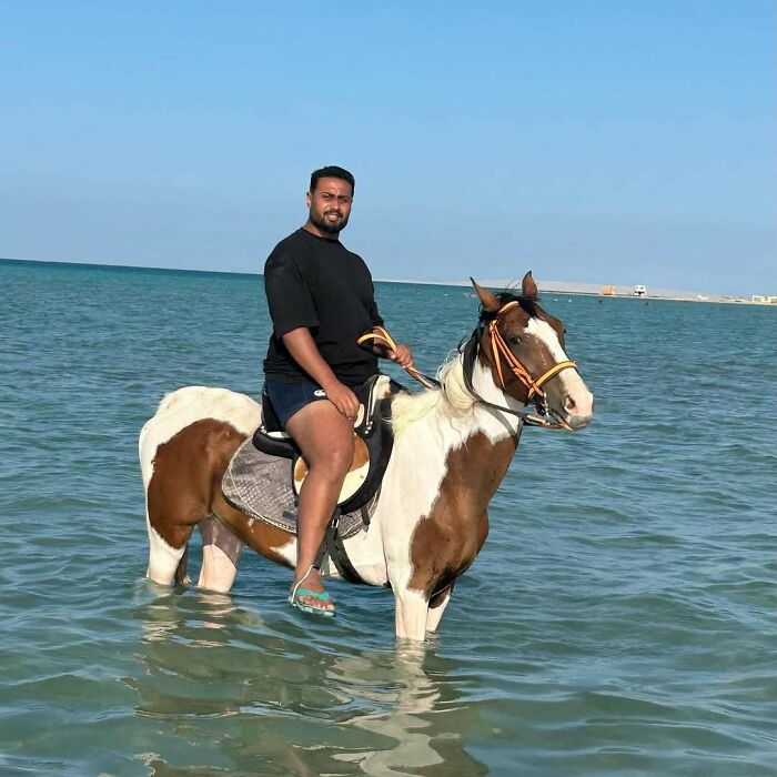 Man in a black shirt and shorts riding a horse standing in shallow ocean water during a vacation.