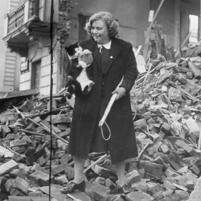 Woman in vintage clothing holding a black and white cat amid rubble in a historic black and white photo of vintage cats.