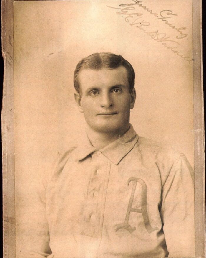 Sepia-toned historical photo of a young man in early 20th century baseball uniform with letter A on chest.