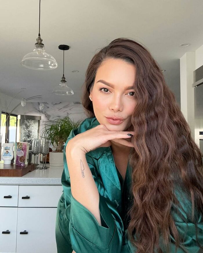 Miss Universe winner posing indoors with long wavy hair and wearing a green satin outfit in a modern kitchen setting.