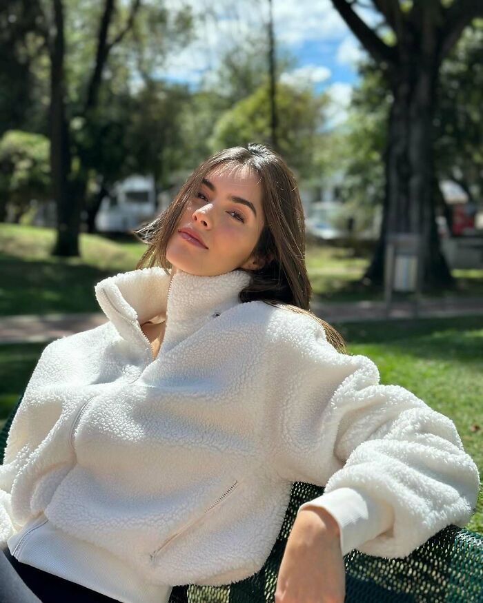 Young woman relaxing on a park bench wearing a white fleece jacket, captured in stunning photos of Miss Universe winners.