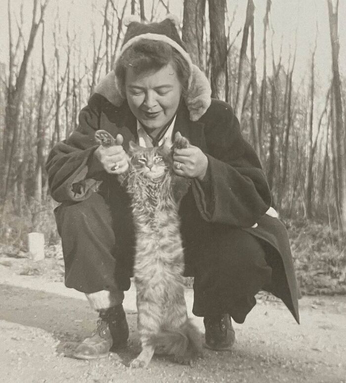 Woman in vintage attire holding up a vintage cat by its front paws outdoors in a bare forest setting.
