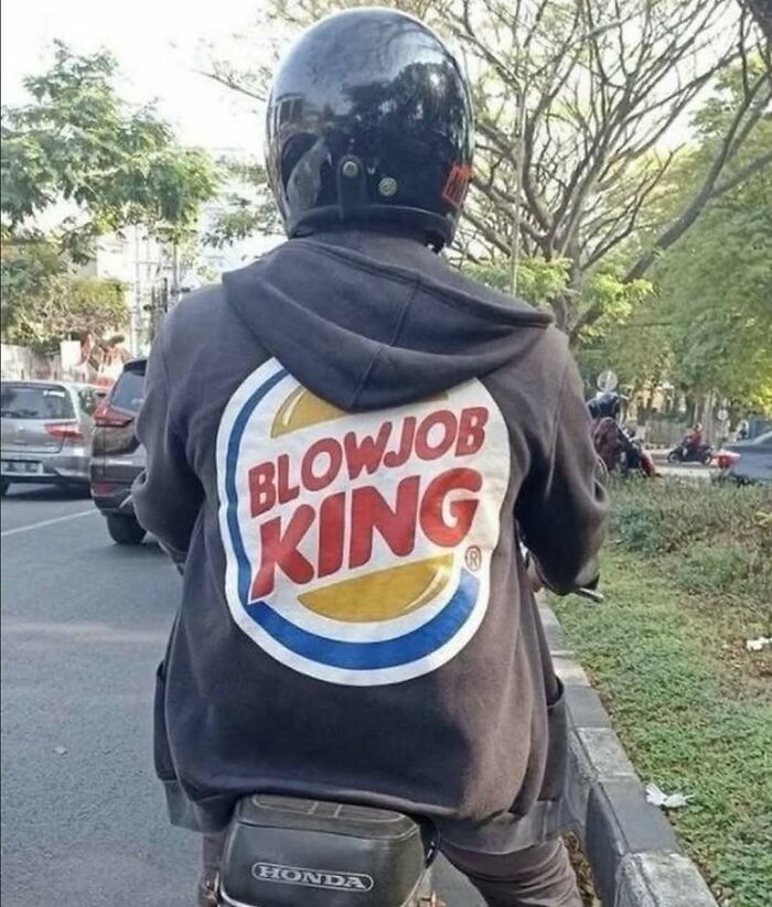 Person riding a motorcycle wearing a hoodie with a humorous cursed image parody logo on the back in an outdoor urban setting.