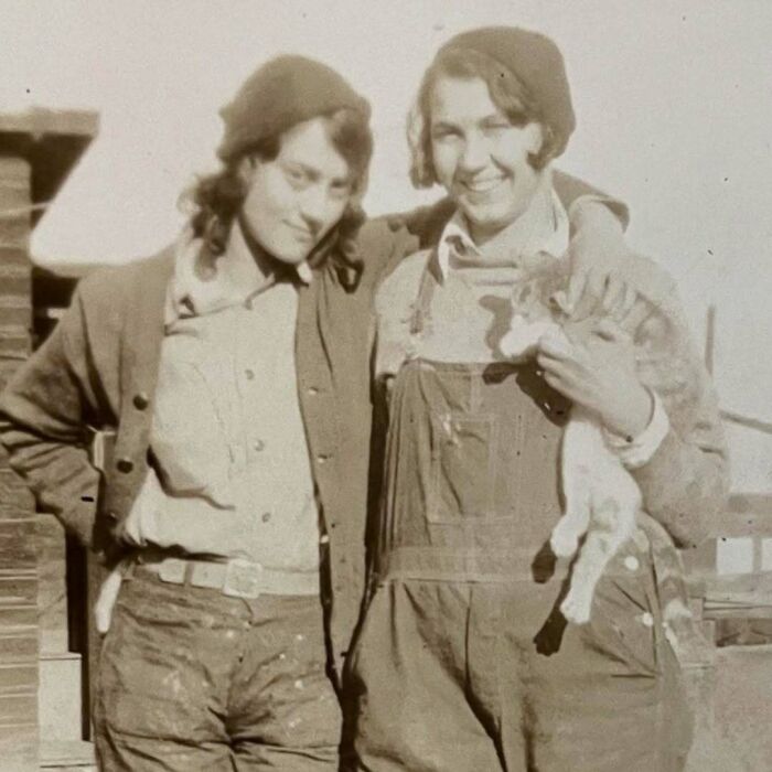Two women in vintage clothing smiling, one holding a cat, showcasing vintage cats in an old photograph.