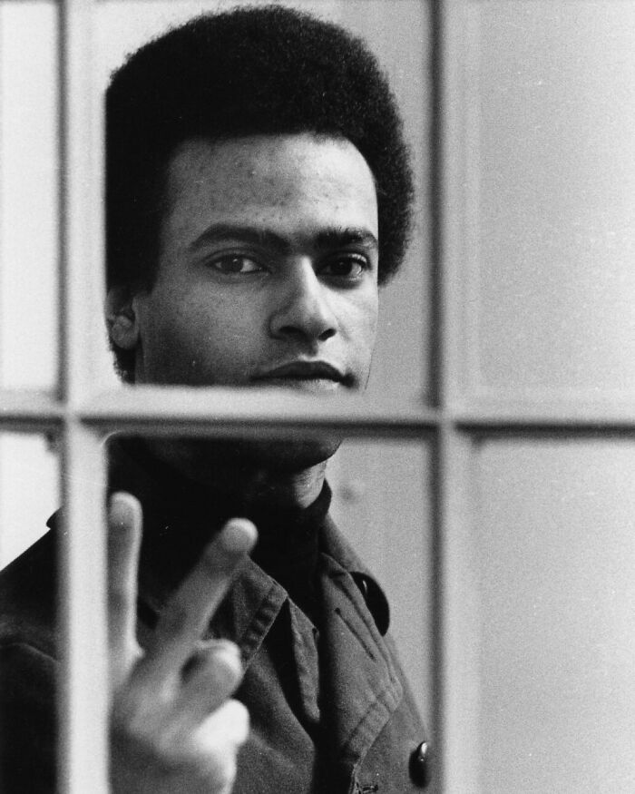Black and white portrait of a man with afro making a peace sign behind a window, showing common folk impact on society.