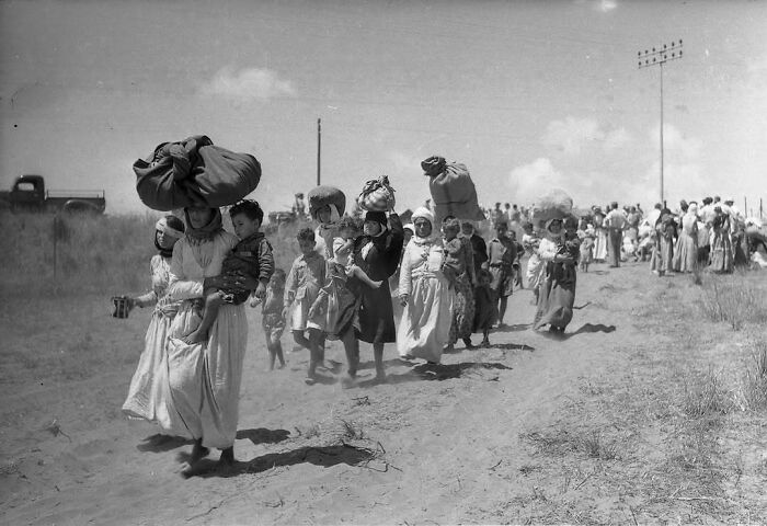 Line of common folk carrying bundles and children, walking along a dirt road under a clear sky in a historic setting.