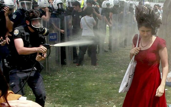 Police officer wearing protective gear spraying pepper spray at a woman in a red dress during a common folk protest.