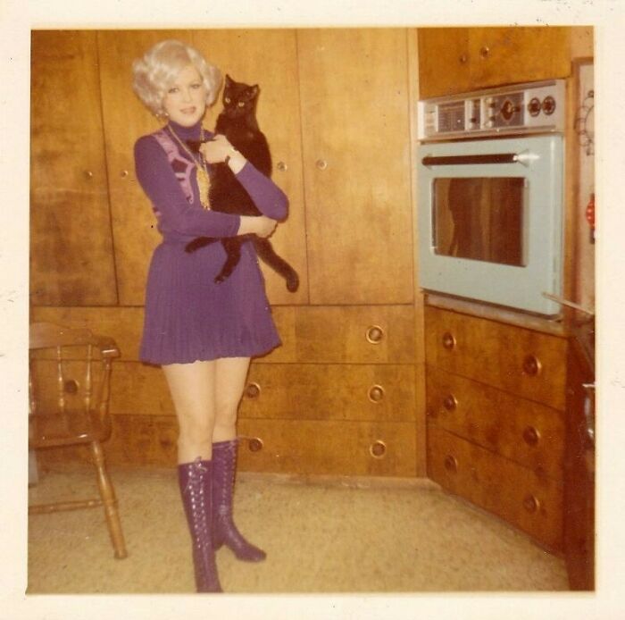 Woman in a vintage purple dress and boots holding a black cat in a retro wooden kitchen, evoking June Cleavers vintage closet.