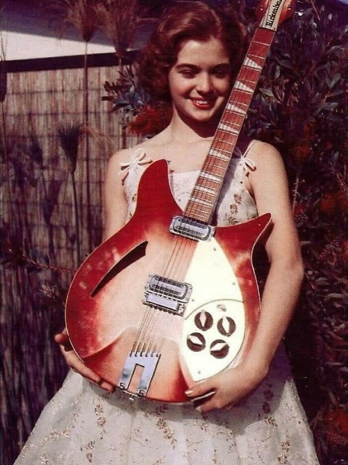 Young woman in vintage dress holding a classic red electric guitar, evoking June Cleavers vintage closet era nostalgia.
