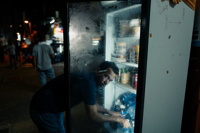 Man bending inside a refrigerator on a dimly lit street, capturing poetic cinematic images of ordinary moments at night.