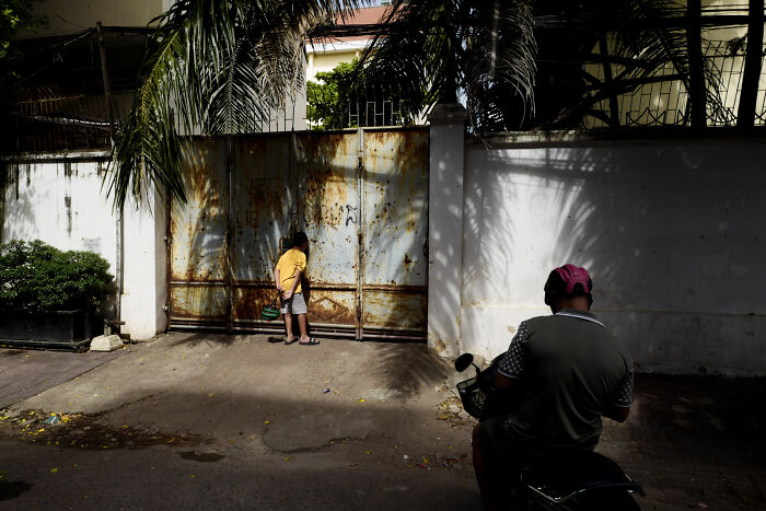 Child in yellow shirt near rusted gate and person on motorcycle in shadowy street, cinematic photographic moment capturing everyday life.
