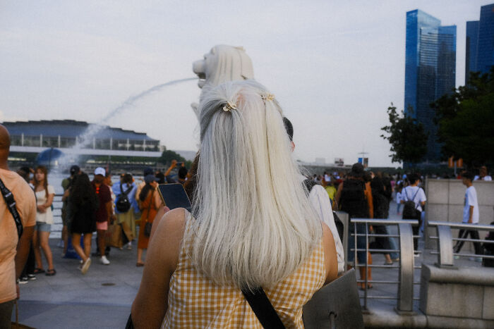 Woman with blonde hair standing near Merlion fountain, captured in a poetic, cinematic image of ordinary moments in a crowded place.
