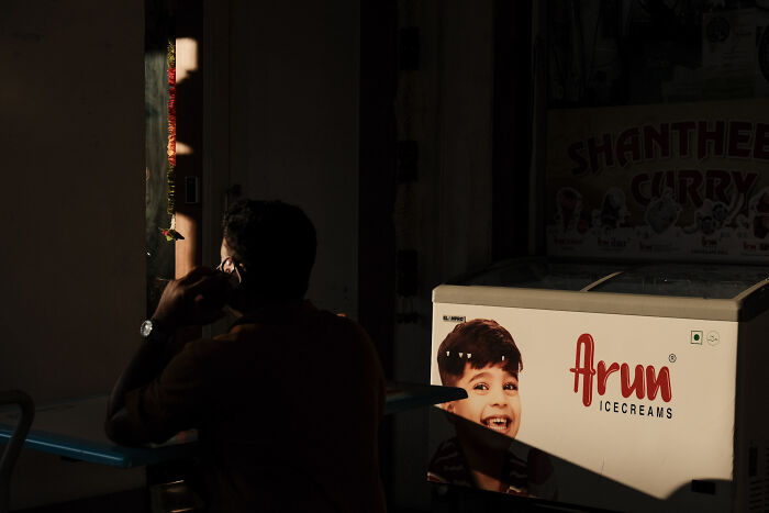 Man sitting in dim light beside an ice cream freezer, captured in a poetic, cinematic style by the photographer.
