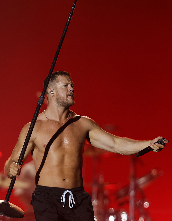 Dan Reynolds shirtless on stage holding microphone stand, performing with red background related to toxic masculinity.