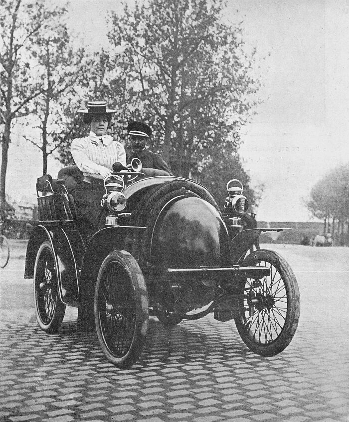 Black and white photo of early wild first cars 100 years ago with two people seated, vintage street setting.