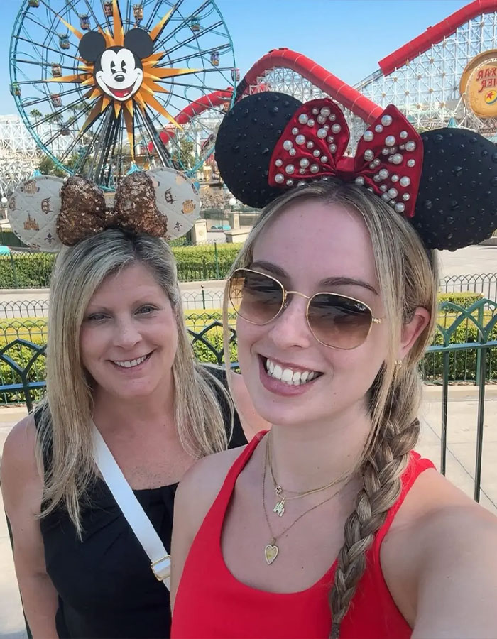 Two women wearing Minnie Mouse ears at Disney World with a Ferris wheel and roller coaster in the background during the day.
