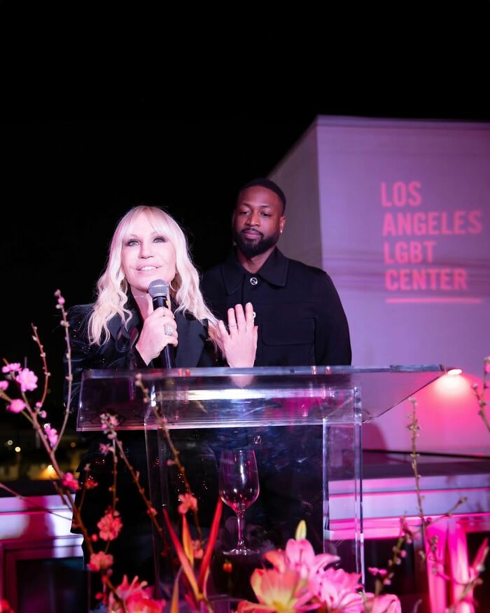 Donatella Versace speaking at an event with a man behind her, showcasing her stunning transformation and youthful appearance.